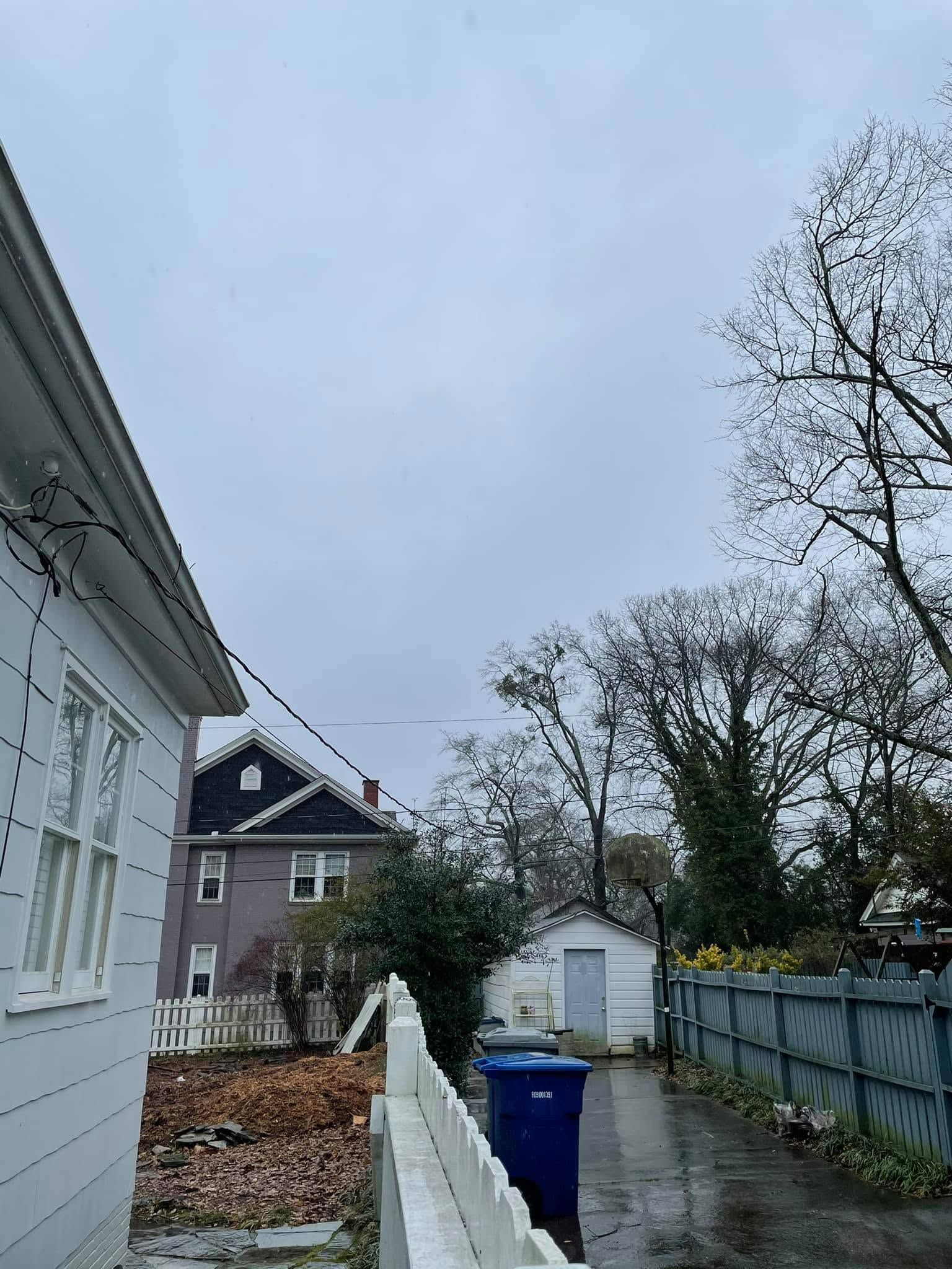 Narrow wet side yard with blue trash bin, white shed, gray fence, and houses under a cloudy sky