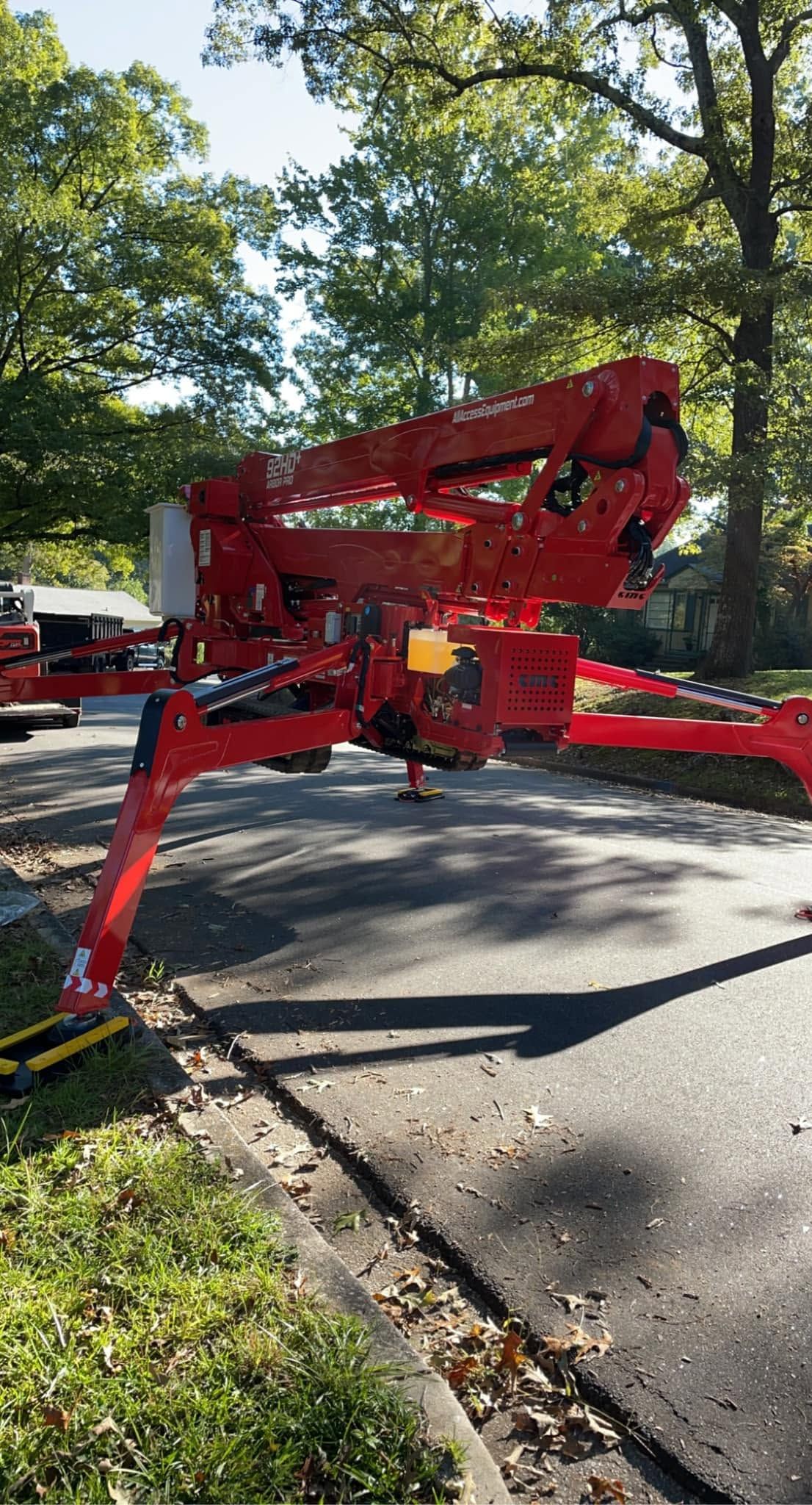 Red construction lift with extended outriggers parked on a tree-lined street in daylight