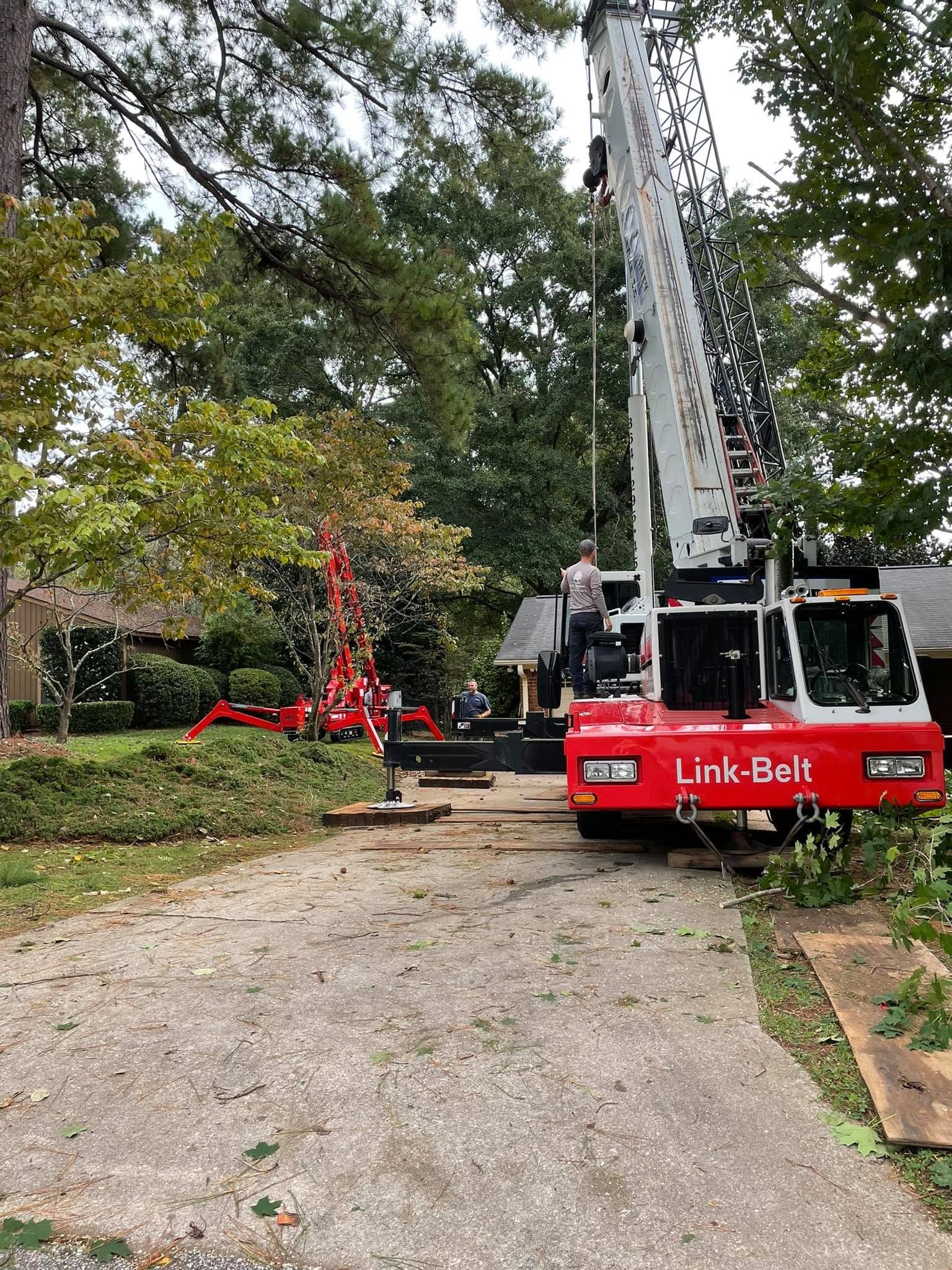 Red Unk-Bolt crane truck on a gravel driveway beside trees, with workers and a red metal lift in the yard