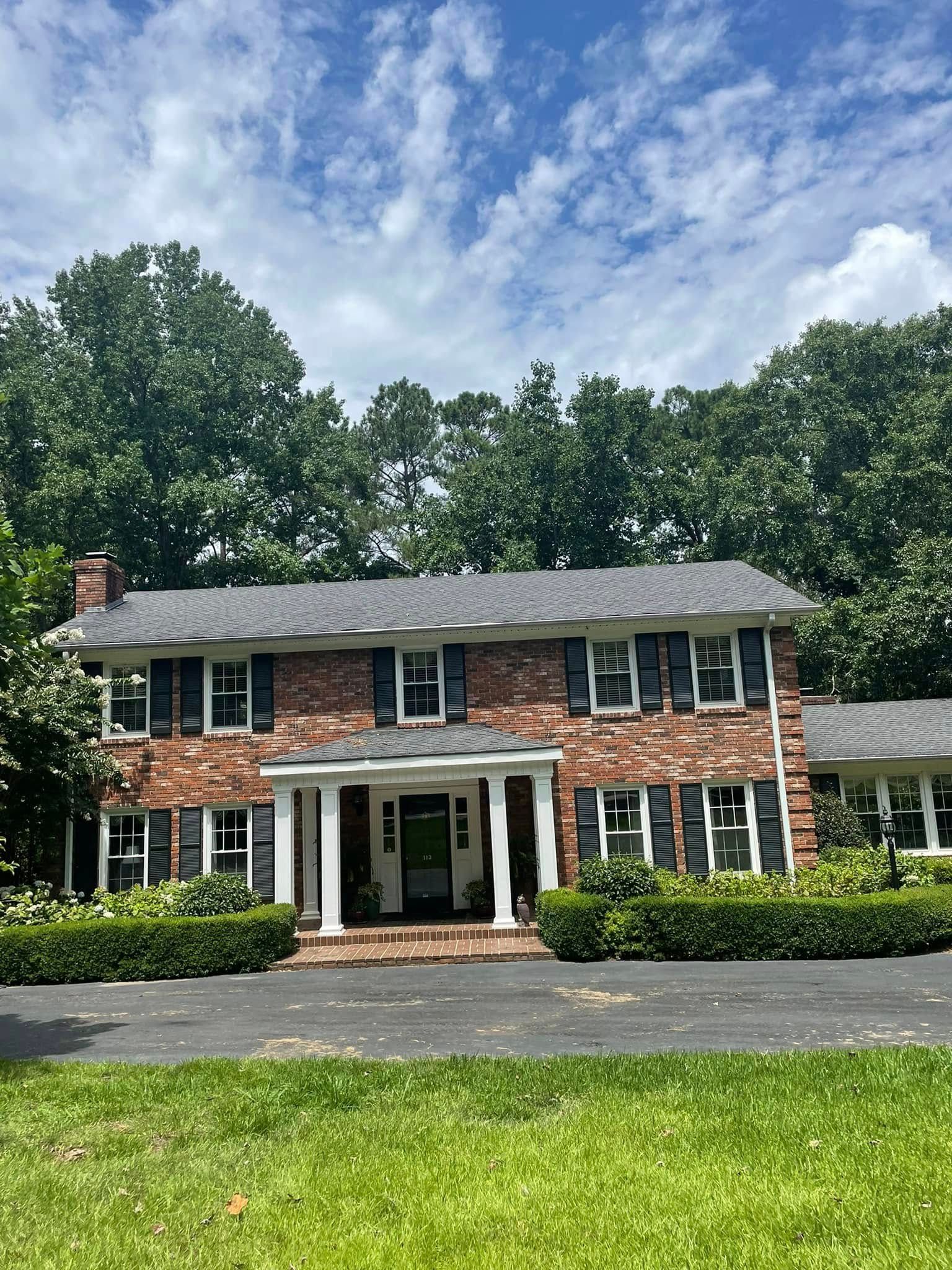 Two-story brick house with white porch, green shutters, and a manicured lawn under a blue sky