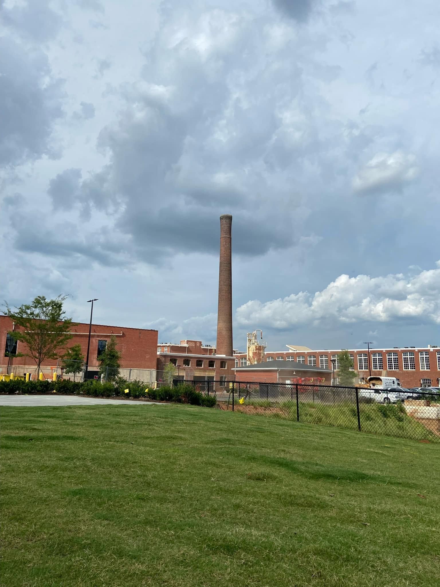 Grassy park overlooking a town skyline with a tall brick smokestack under cloudy skies