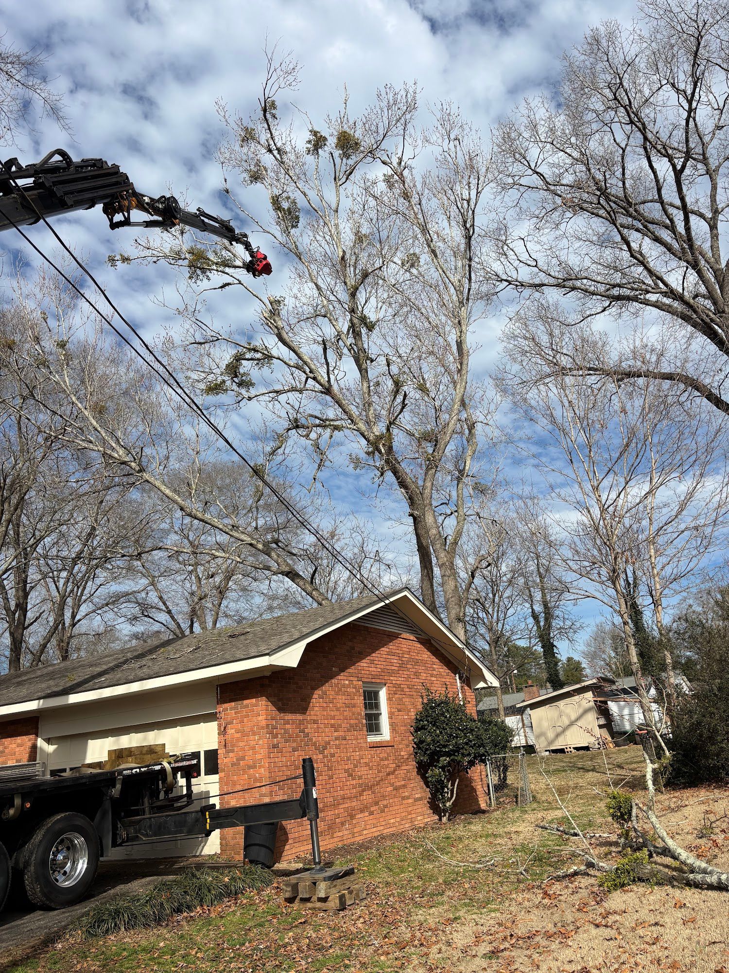 Tree-trimming truck with extended crane beside a brick house in a yard with leafless trees