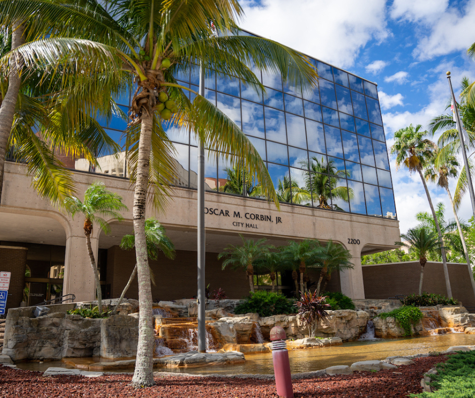 Oscar M. Corbitt Jr. City Hall building with reflective glass facade, palm trees, and water feature.