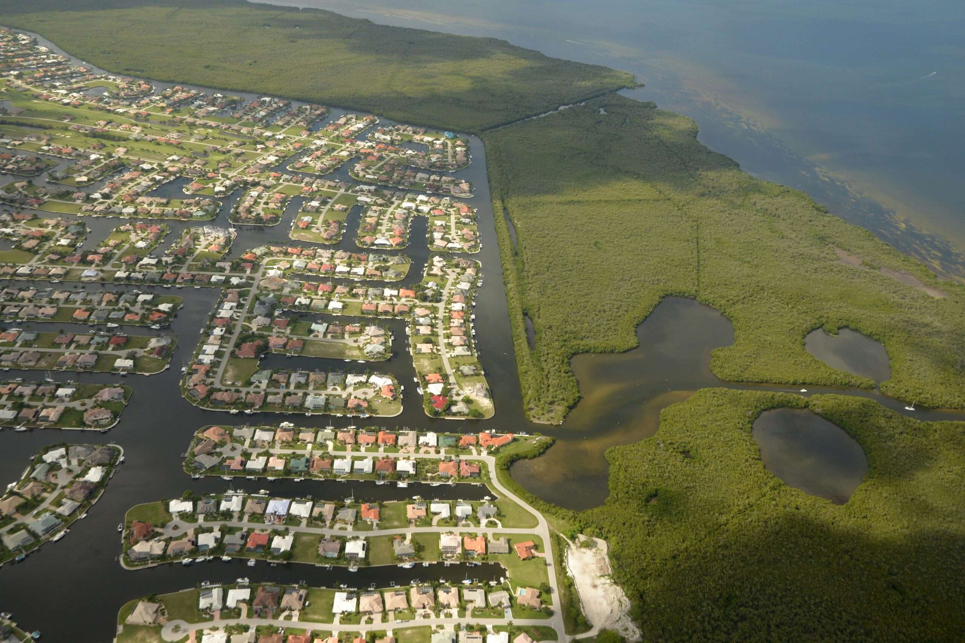Aerial view of a coastal neighborhood with canals bordering a forested area and open water.