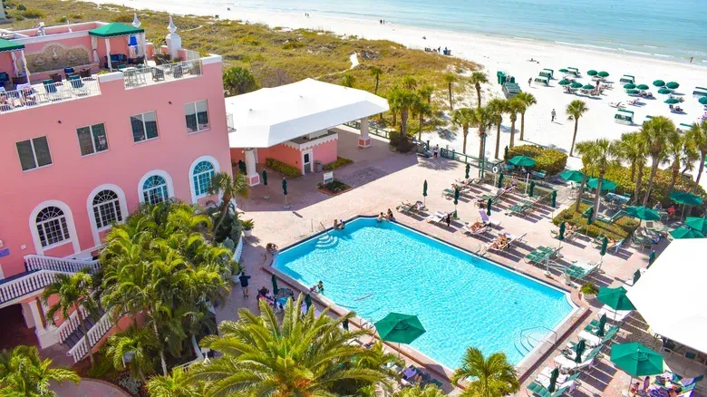 Pink hotel next to a beach with a blue pool. People relax by the pool and on the beach.