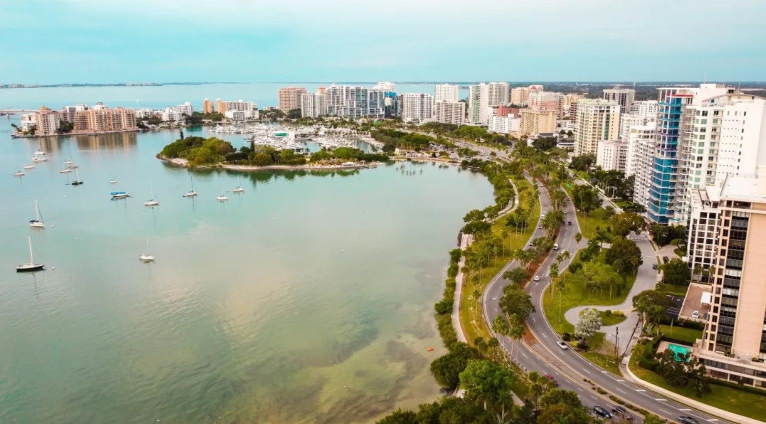 Aerial view of city waterfront with buildings, a marina, boats, and a coastal road.