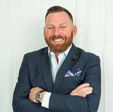 Man with a beard in a blue suit, arms crossed, wearing a watch, smiling.