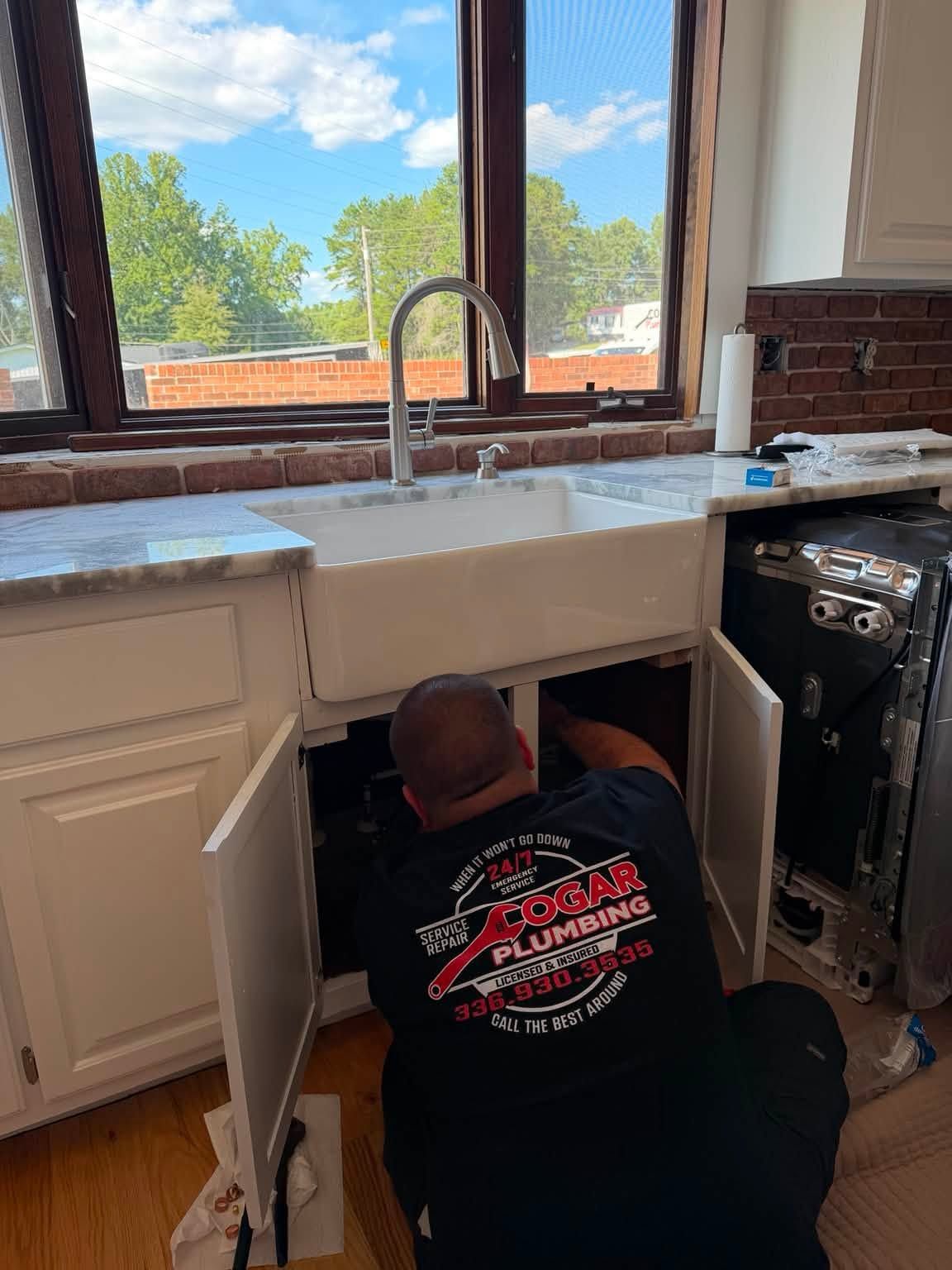 Worker installing a kitchen sink beneath a window, with cabinets open and tools on the counter.
