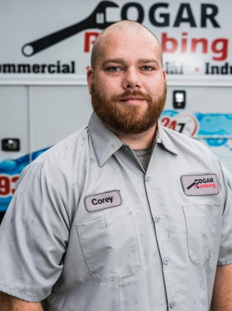 Portrait of a bearded man in a gray work shirt standing in front of a plumbing company sign