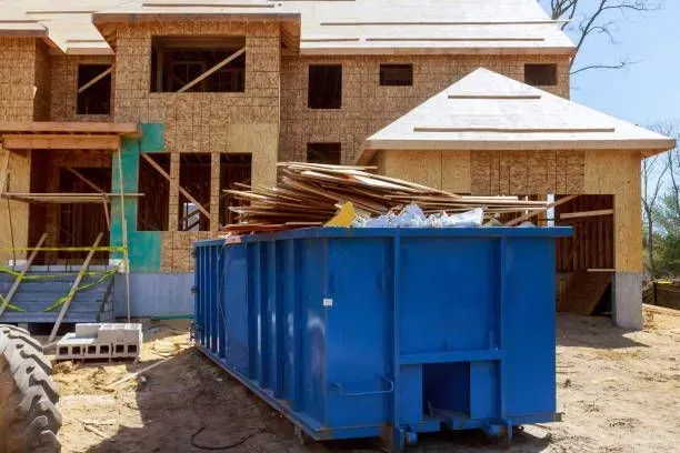 Blue dumpster overflowing with wood debris, construction site in background, unfinished house.