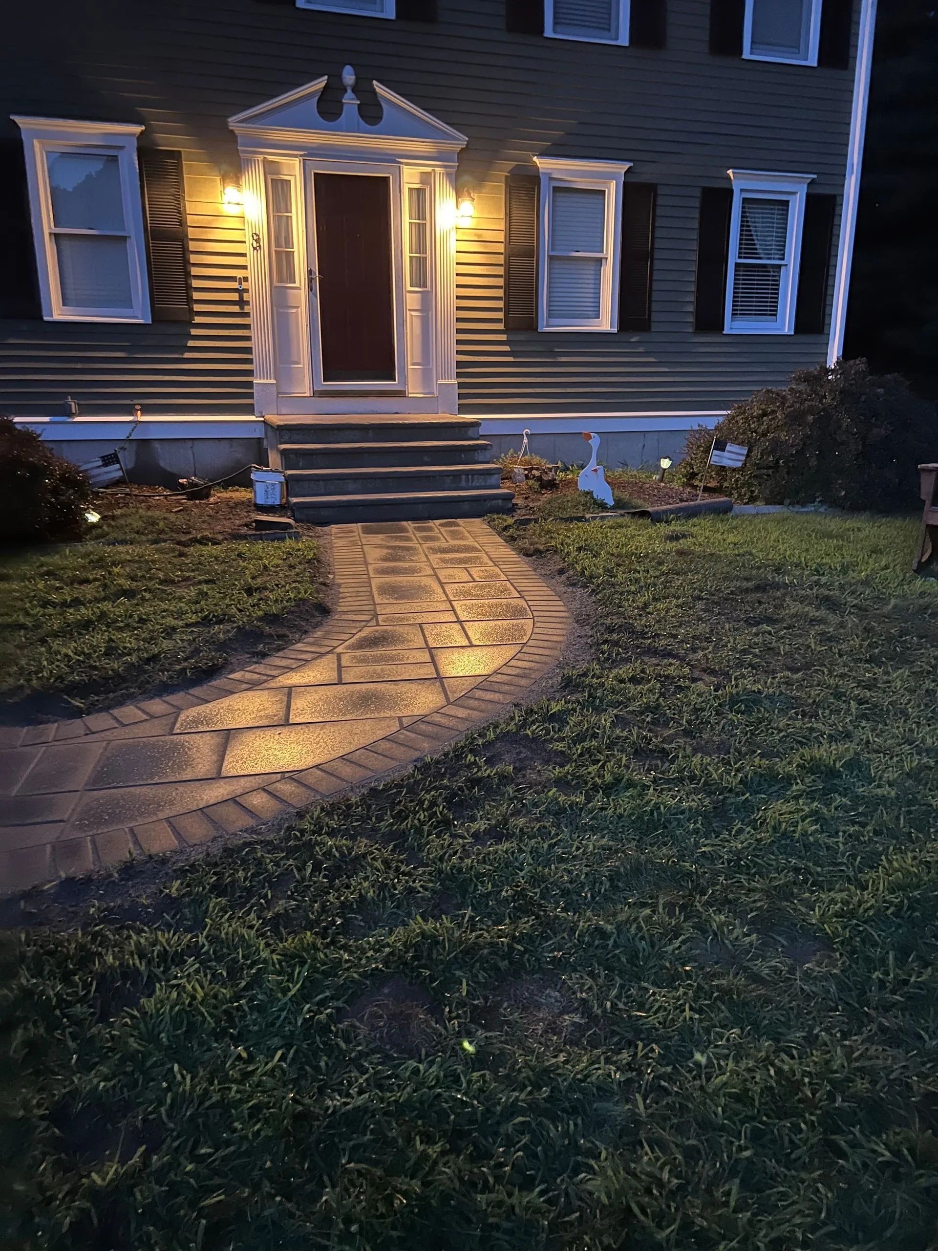 Lit walkway leads to a house with a dark door and glowing lights. Green grass surrounds the path.