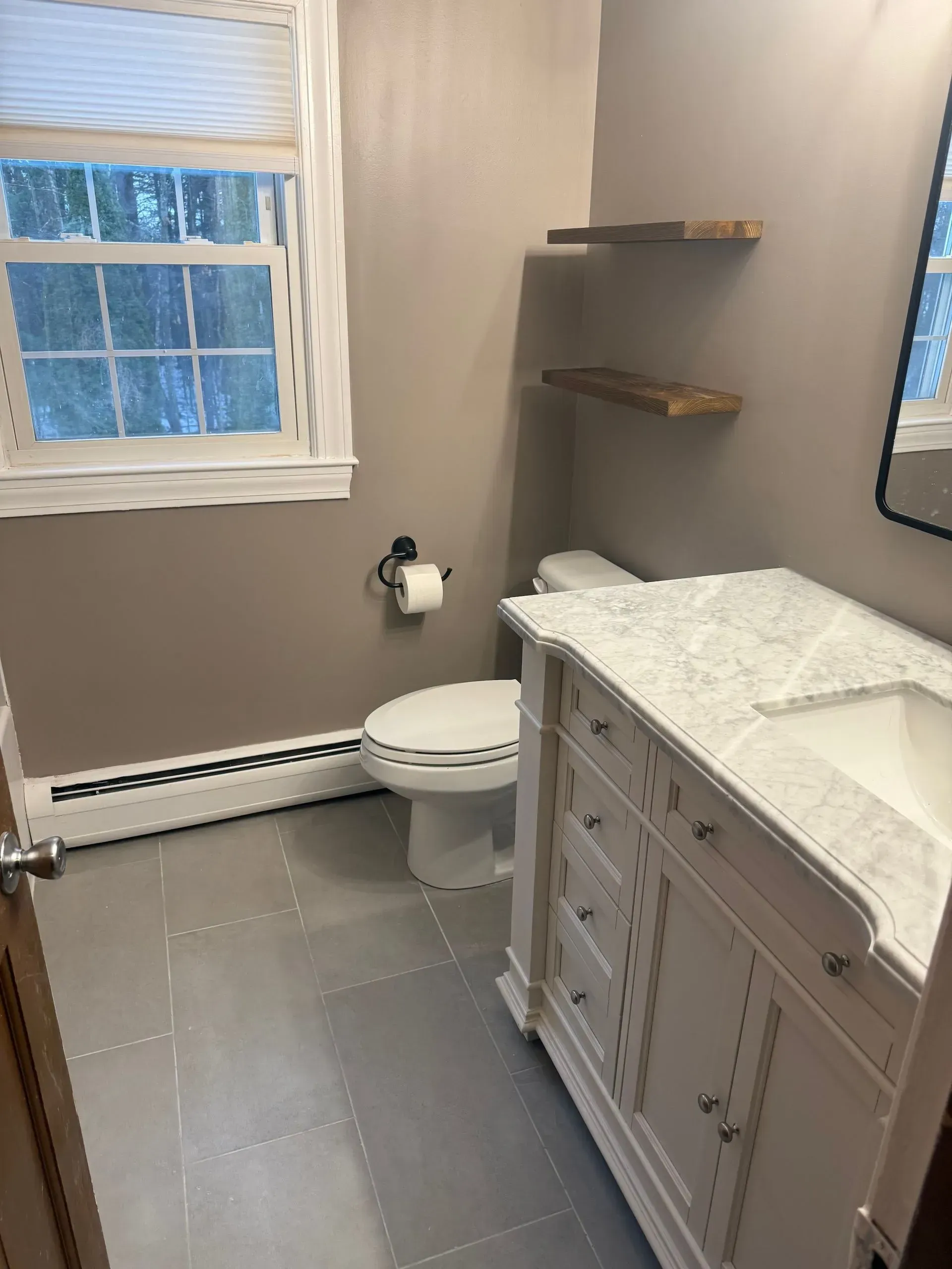 Bathroom with gray tile, white vanity, toilet, window, and corner shelves.