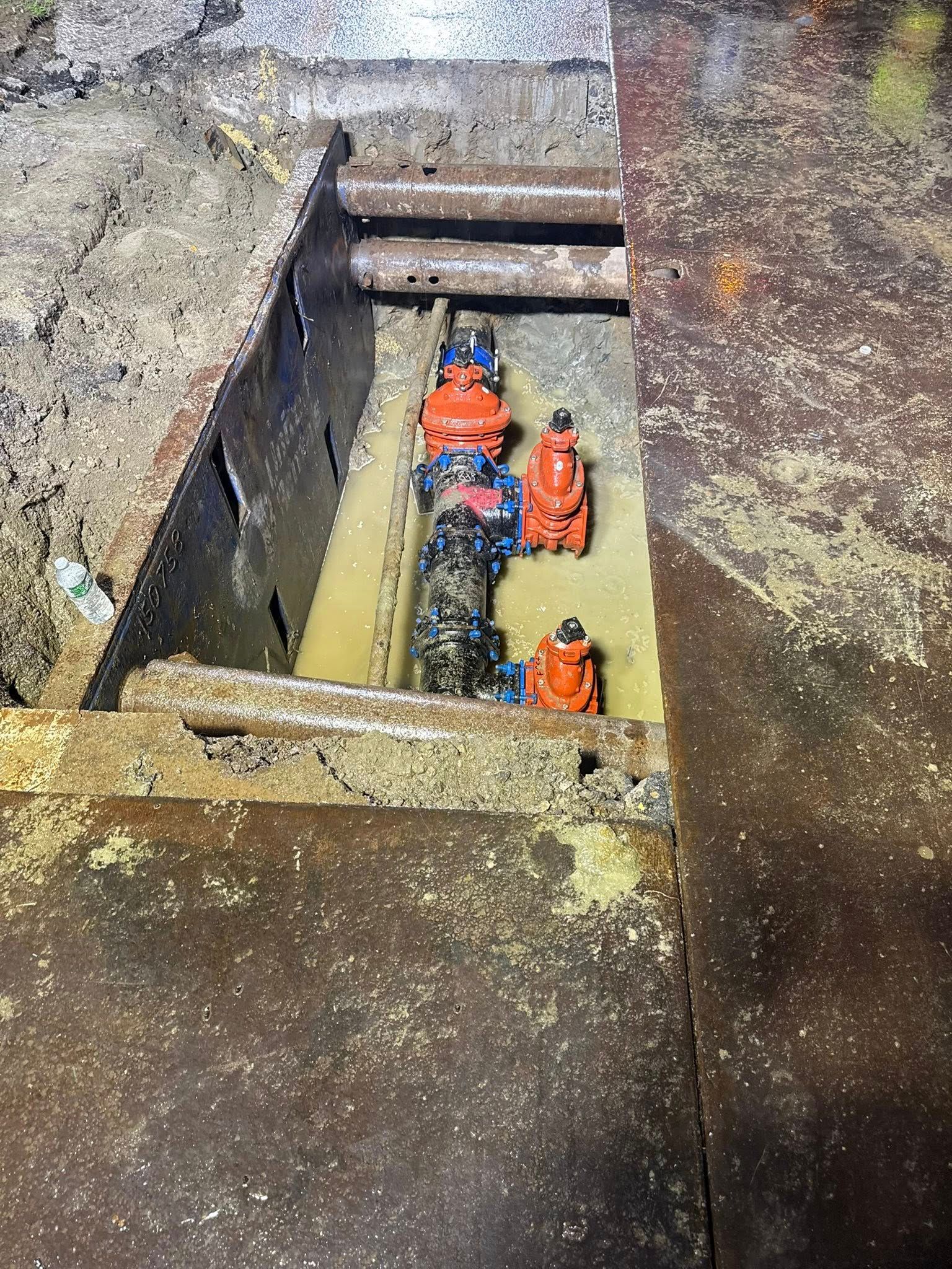 Metal pit revealing pipes and valves, including orange and blue components, in a muddy environment.