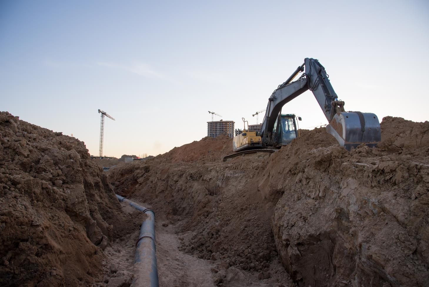 Excavator digging a trench for a pipe on a construction site, with buildings and cranes in the distance.