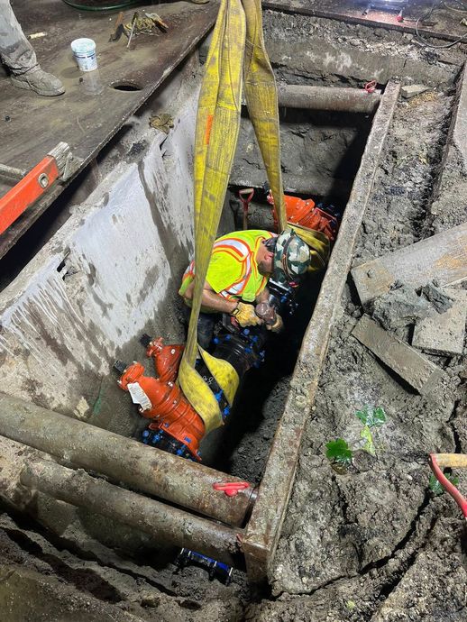 Worker in trench being lifted by yellow straps. Yellow vest, hard hat. Dark trench, concrete, and soil.