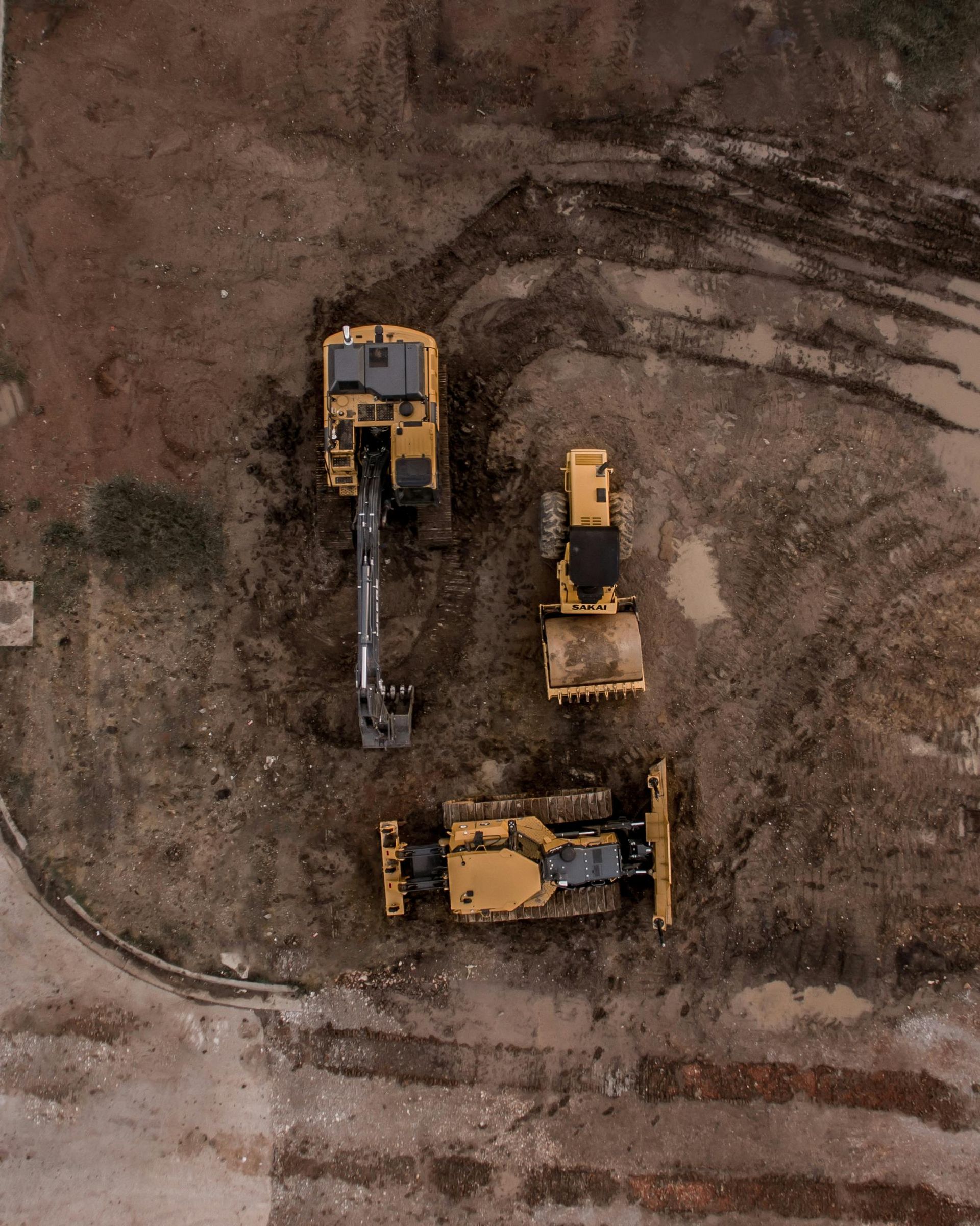 Aerial view of three yellow construction vehicles on a muddy construction site, likely earth moving.