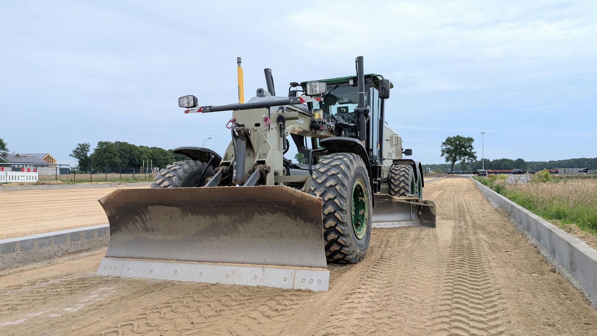 A large, green and gray grader levels sand on a construction site next to a concrete curb.