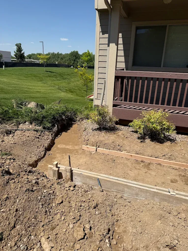 A ground-level view of a residential construction site showing wooden formwork for a new concrete walkway near a deck.
