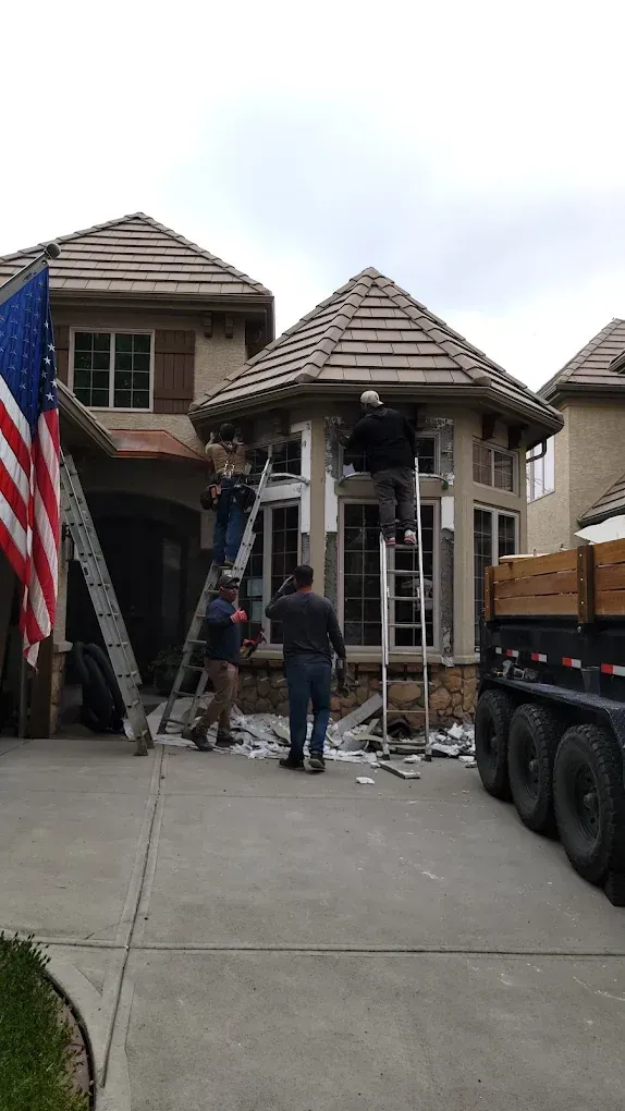 Workers on ladders and the ground perform exterior home renovations in front of a parked trailer and an American flag.