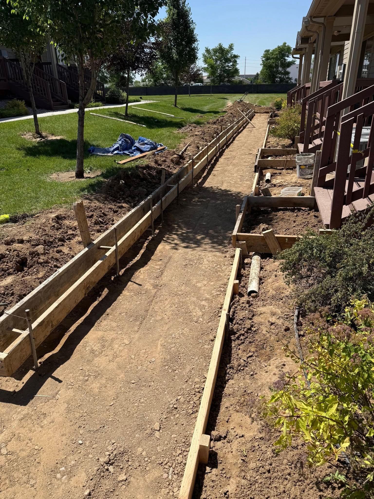 A narrow dirt path lined with wooden forms for a new concrete sidewalk, located beside a building and lawn.