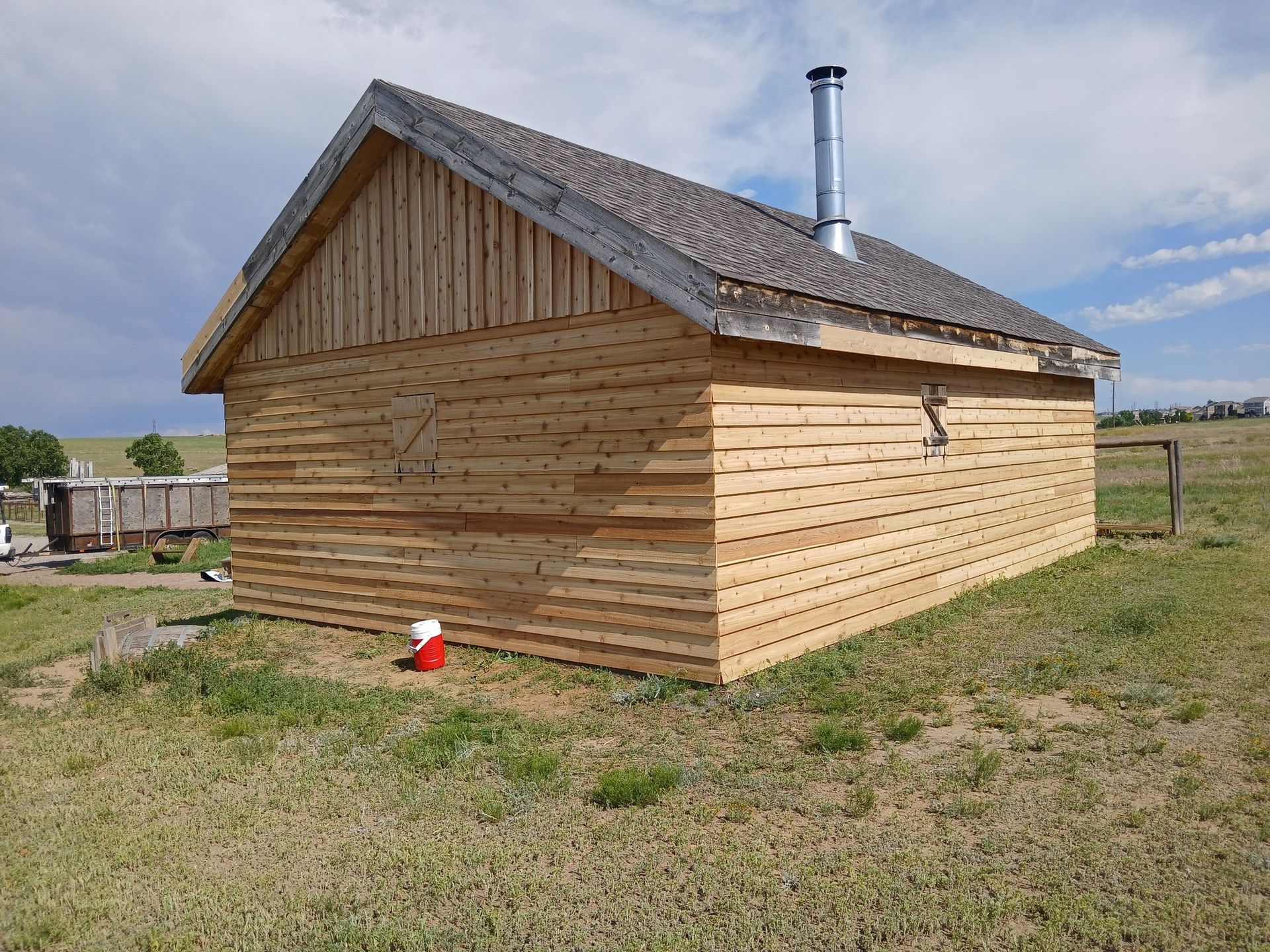 A small wooden cabin with horizontal siding and a metal chimney stands in a grassy, open field under a blue sky.