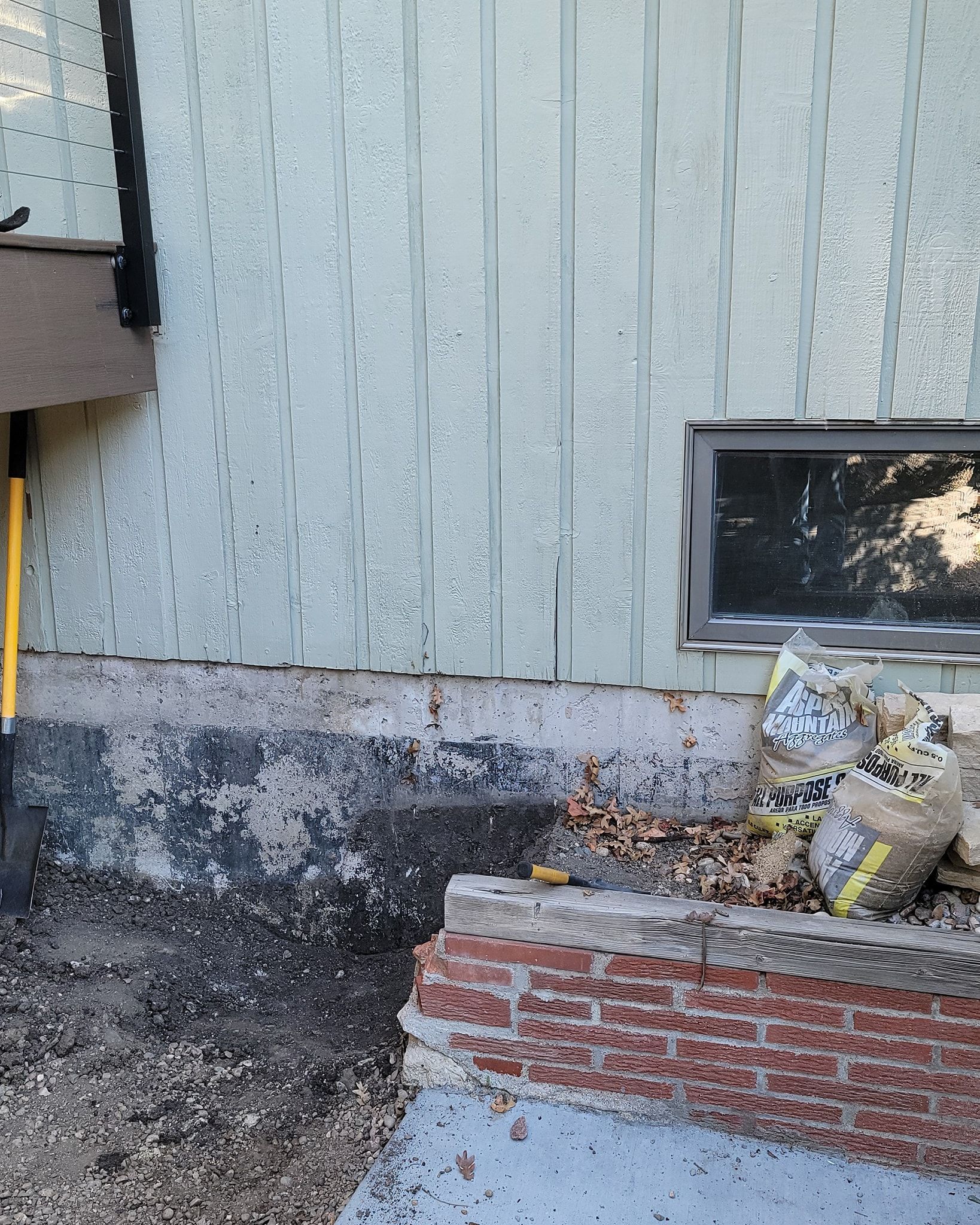Light green vertical siding above a dark concrete foundation and a low red brick wall with two bags of soil near a window.