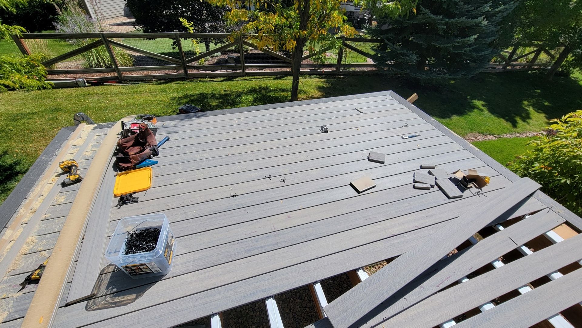 A deck under construction with gray composite planks being installed, scattered tools, and a backyard background.