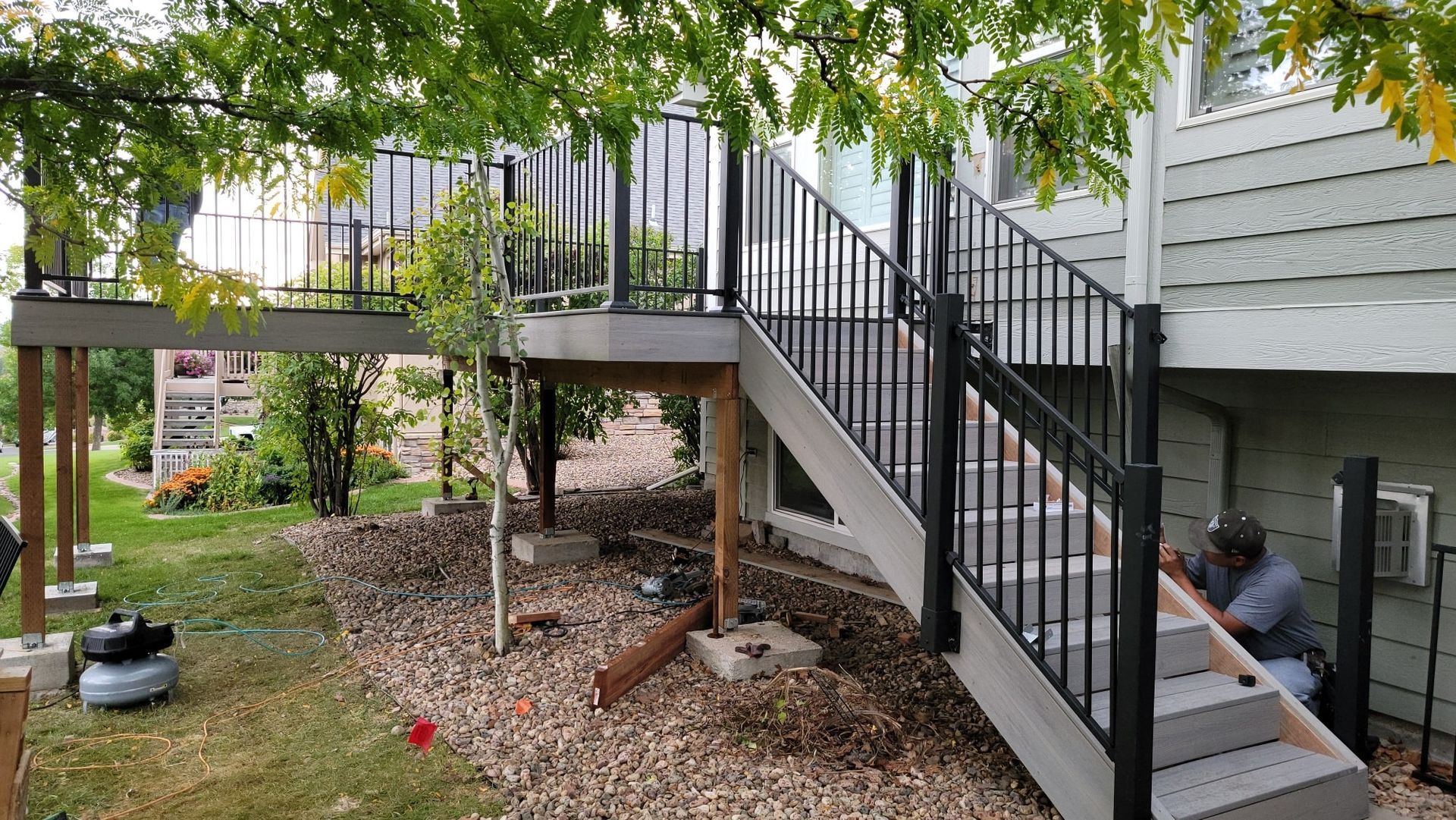 A person in a gray shirt works on the handrail of a new gray wooden staircase attached to a residential deck.