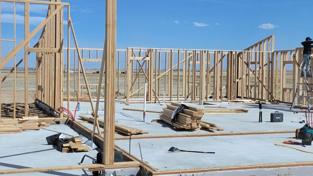 A person stands on a ladder working on the wooden wall framing of a house under construction on a concrete foundation.