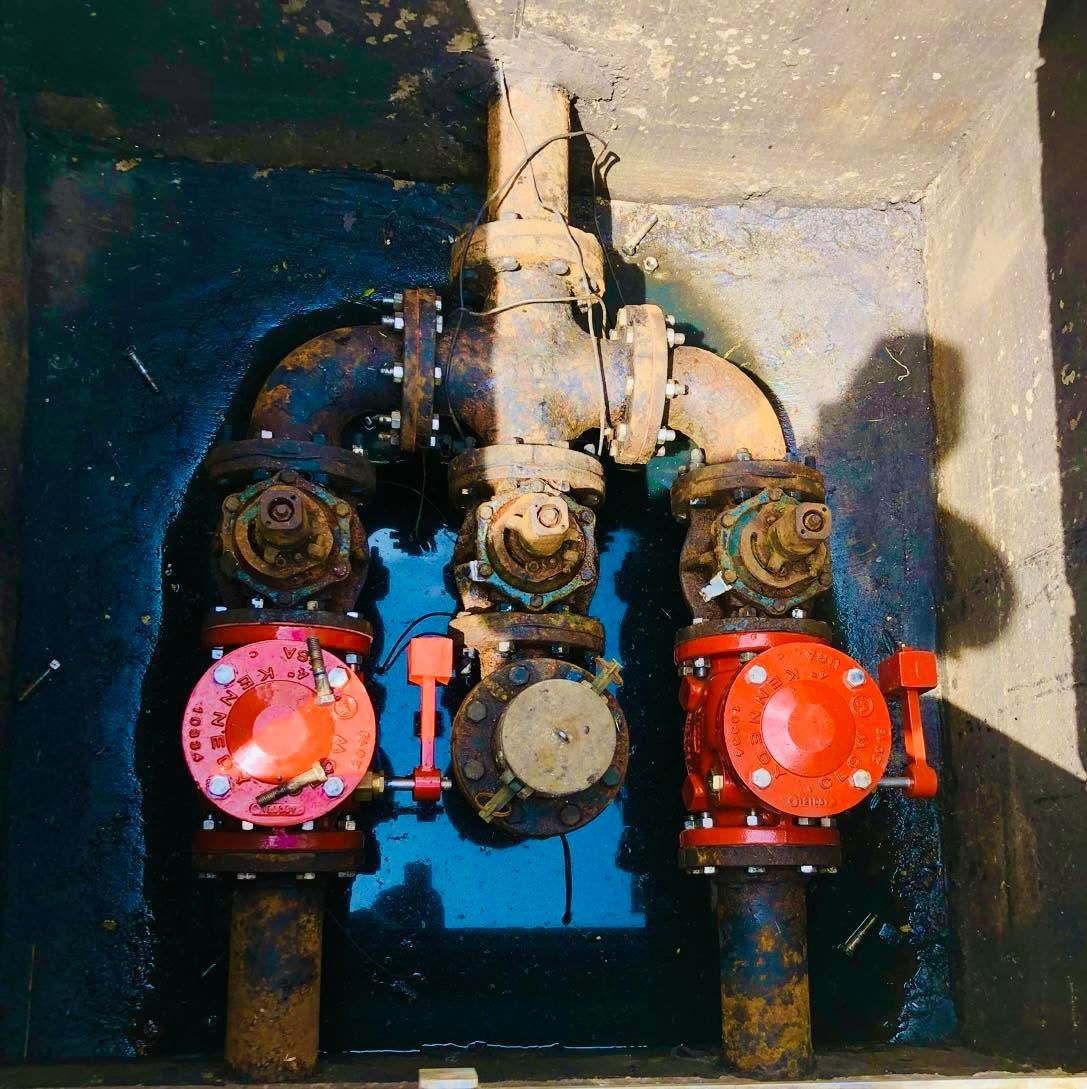 Valves and pipes in a small utility pit, with red and blue wheel handles on a concrete wall.