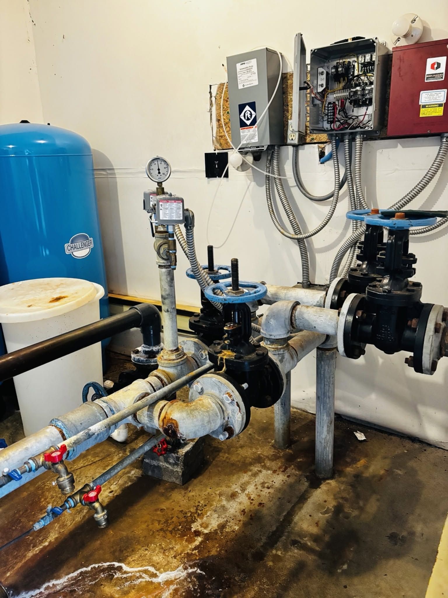 Industrial pump and piping system in a utility room, with blue tank, gauges, and wall-mounted controls.