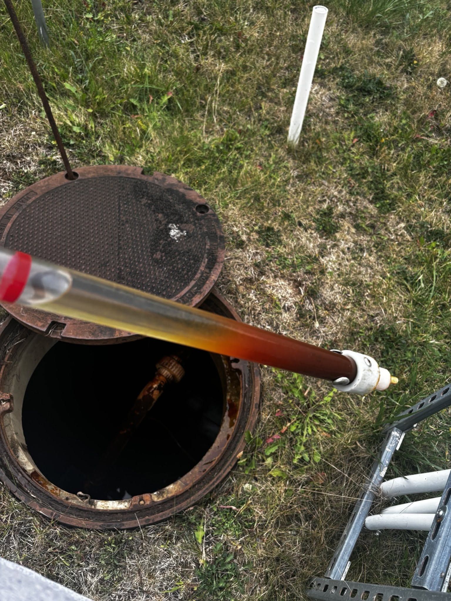Worker lowering a clear tube into an open manhole beside a ladder and grass-covered ground