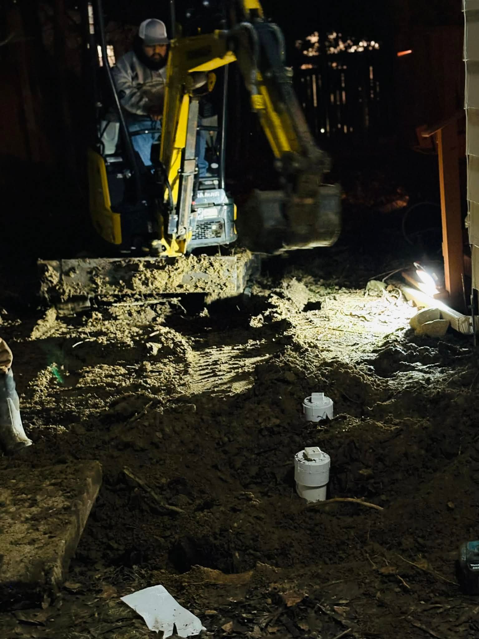 Worker operates an excavator at night in a muddy trench, with bright work lights and sparks nearby.