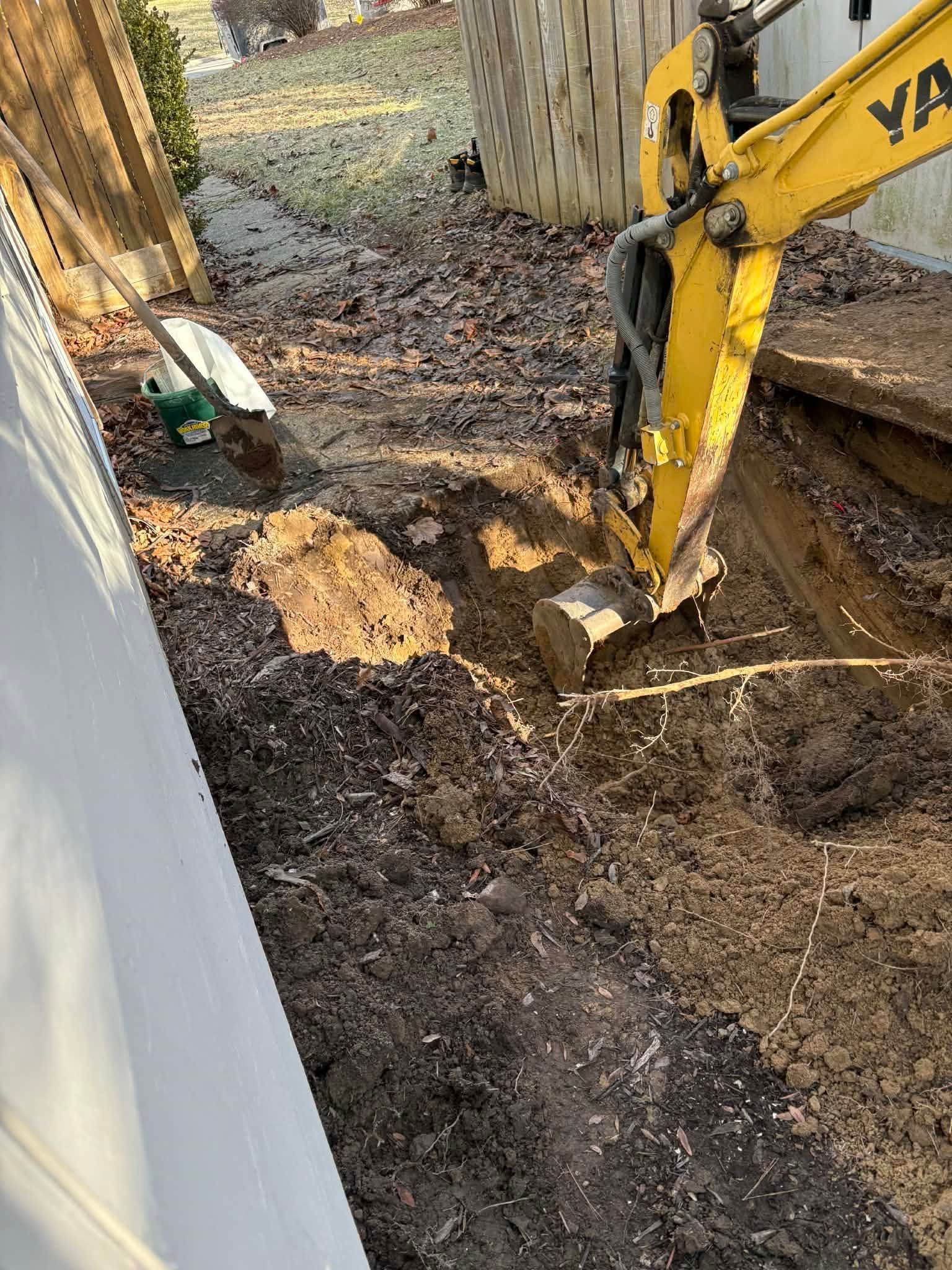 Yellow excavator digging a trench beside a building in a muddy yard