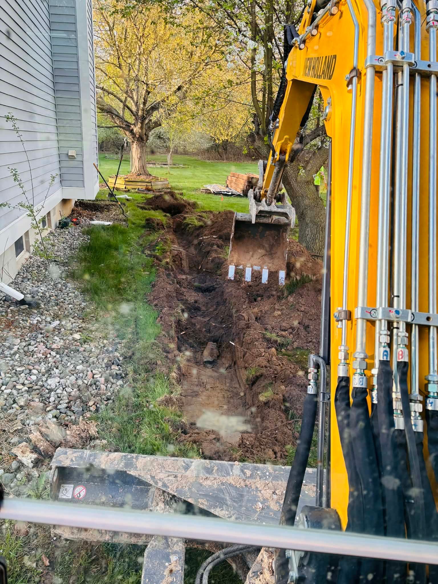 Yellow excavator digging a trench beside a house, with dirt and gravel along the foundation.
