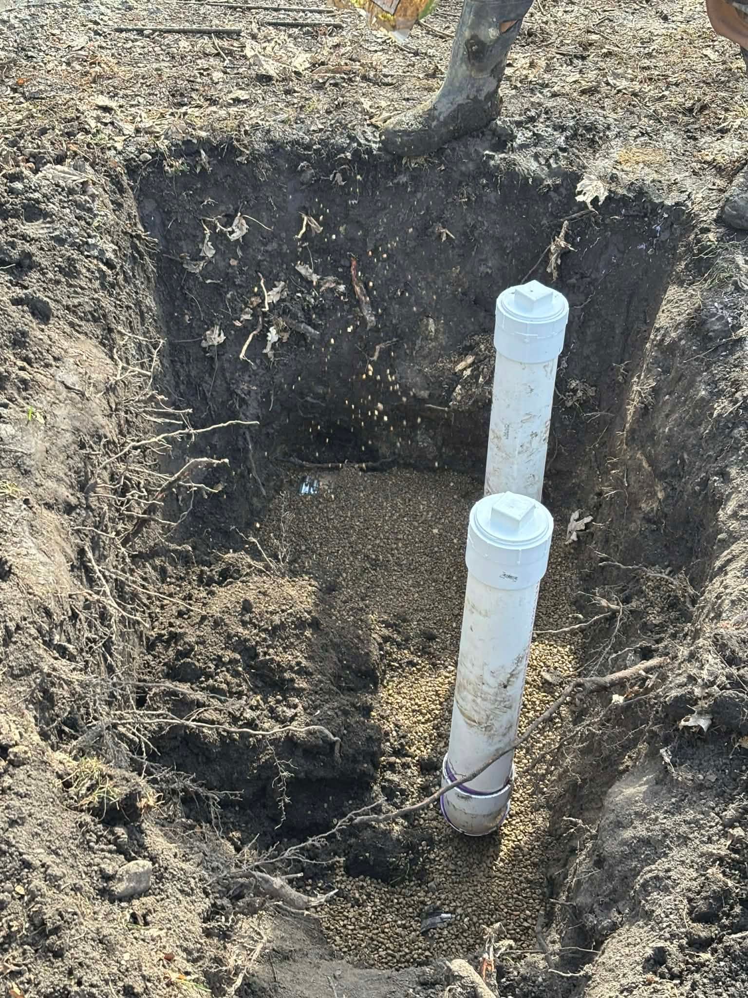 Open trench with a white PVC pipe standing upright in dirt and gravel.