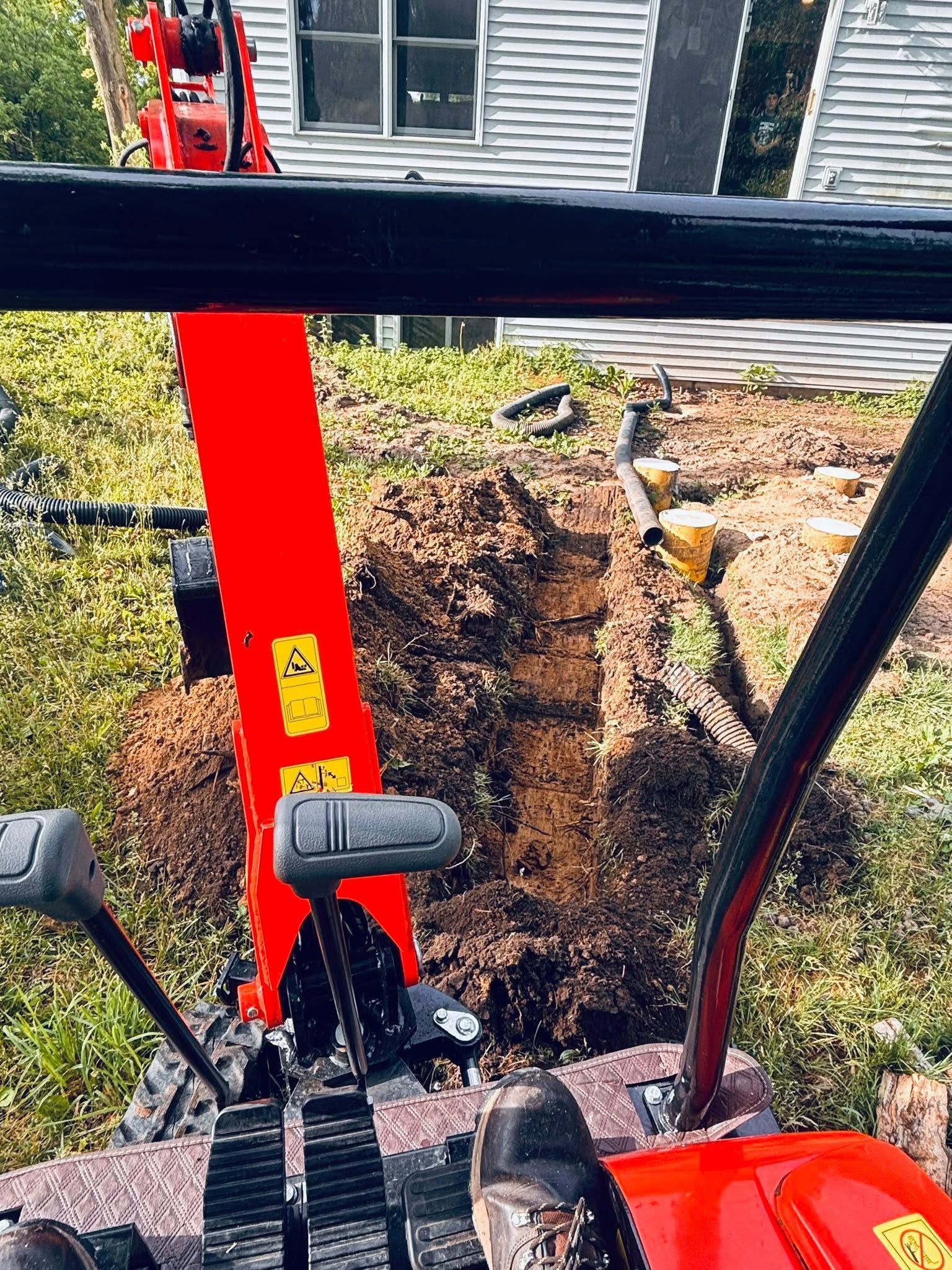 Red mini excavator digging in a grassy yard beside a house foundation.