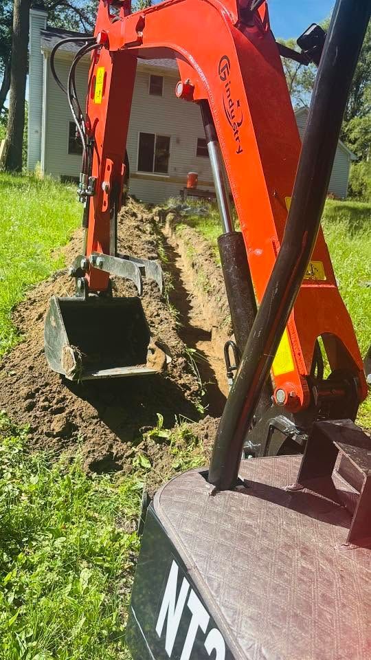 Orange excavator digging a trench beside a grassy yard and house.