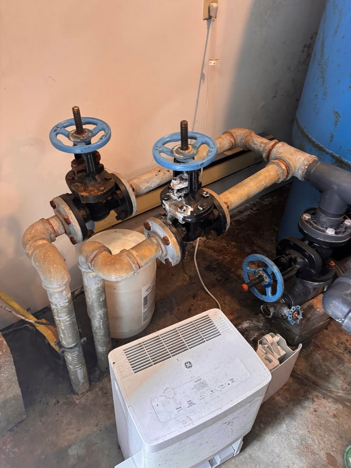Industrial pipes and blue valve wheels beside a white floor fan in a basement utility area