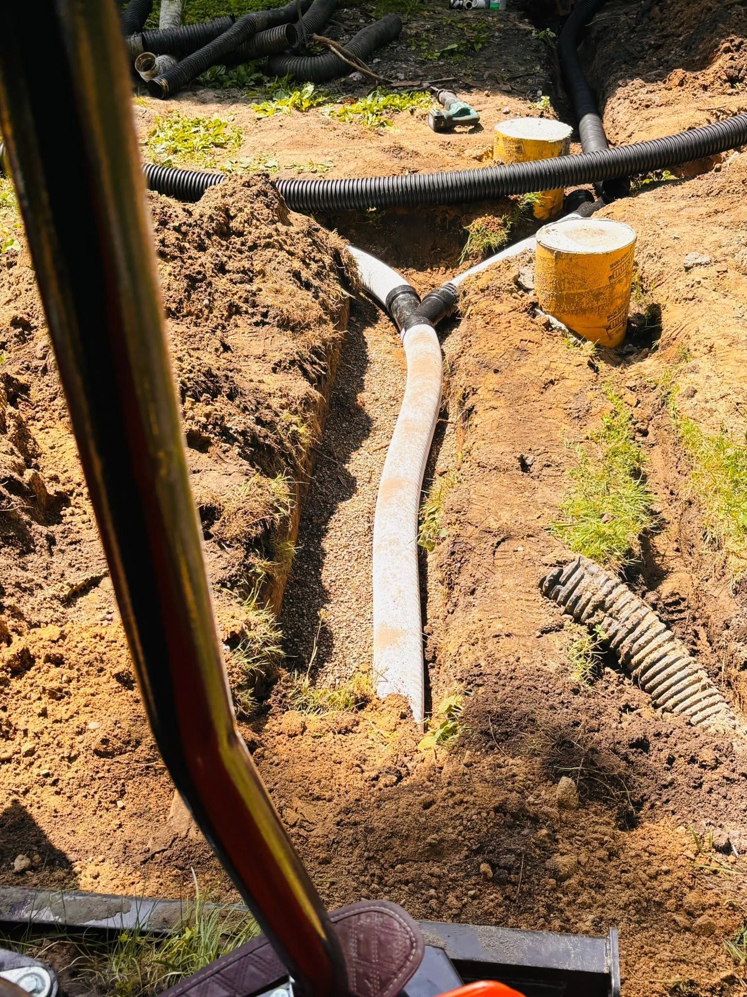 Trench with exposed utility pipe and black cables in sandy soil, with a yellow cap nearby.