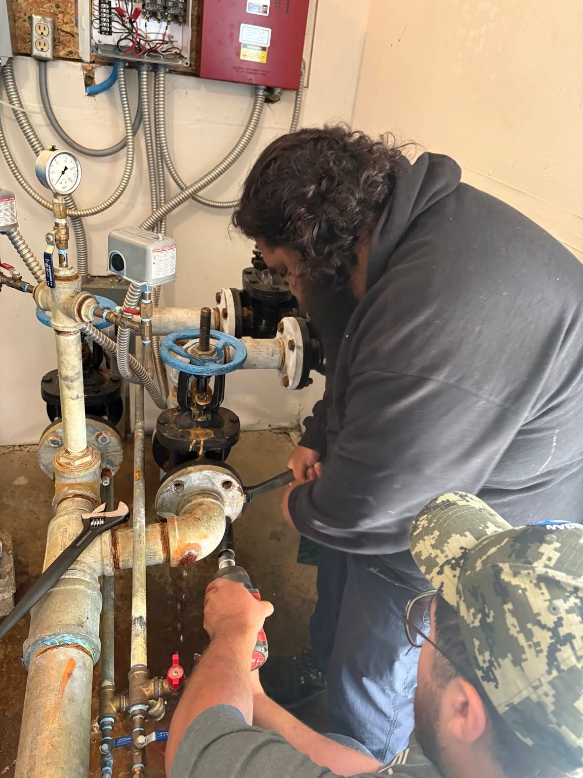 Worker inspecting pipes and valves in a utility room, with tools and gauges on the wall.