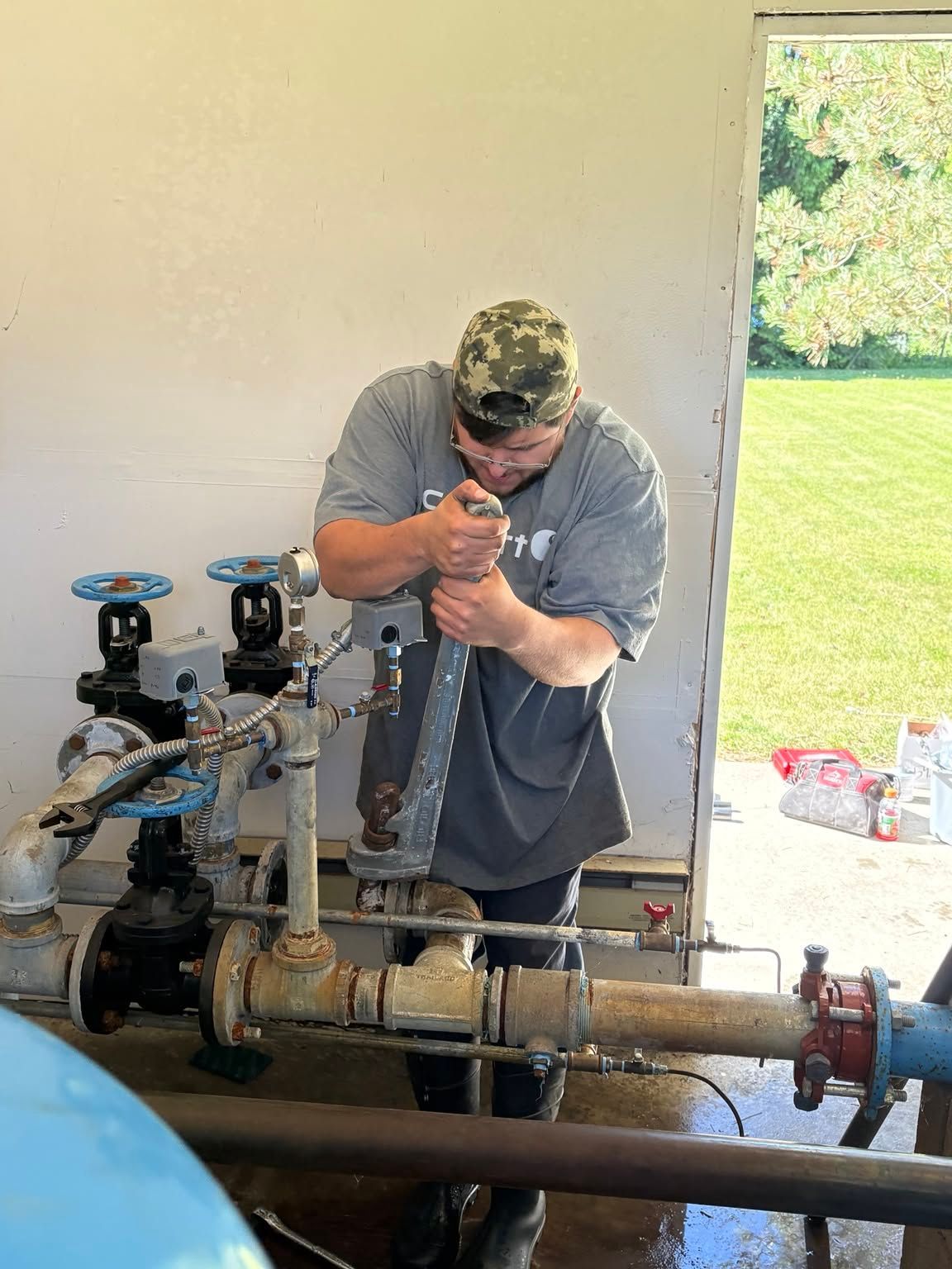 Worker tightening a large pipe valve indoors, with plumbing equipment and a grassy yard visible outside.