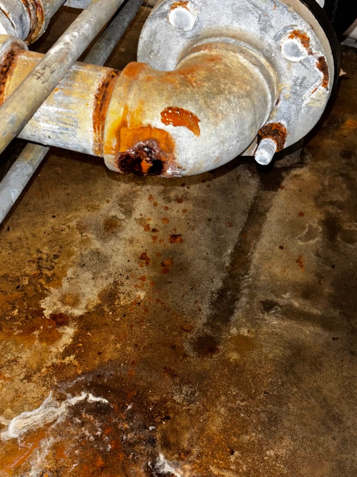 Rusty metal pipe connection with orange corrosion and a damp, stained floor nearby