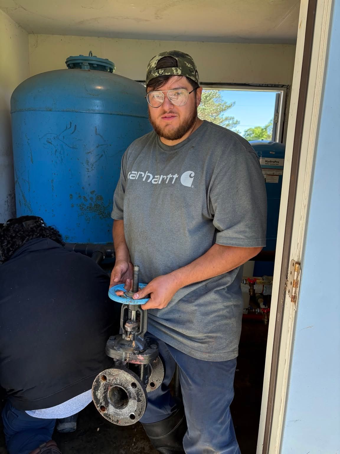 Man in gray shirt holding a metal valve in a small utility room with a blue tank behind him