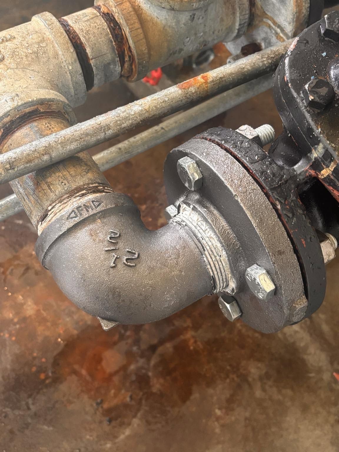 Close-up of a rusted metal pipe elbow with a bolted flange and valve in an industrial setting