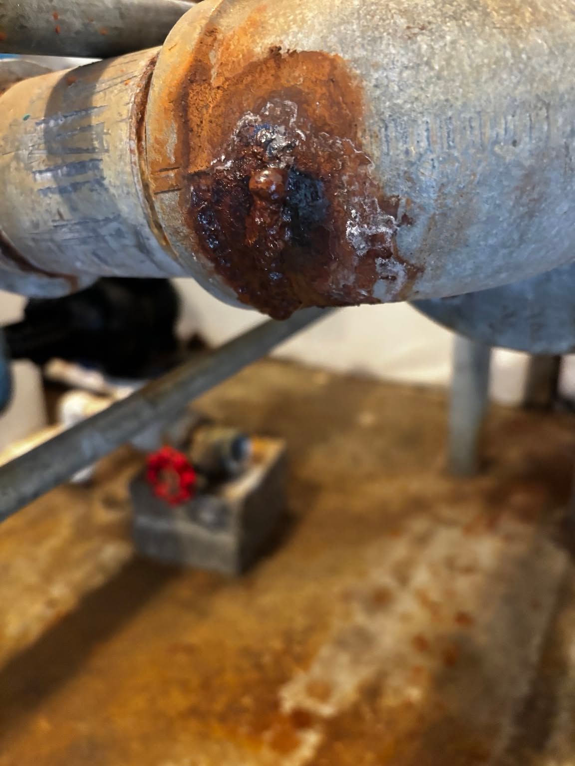 Close-up of a rusty, corroded metal pipe joint with a rusted valve in the background