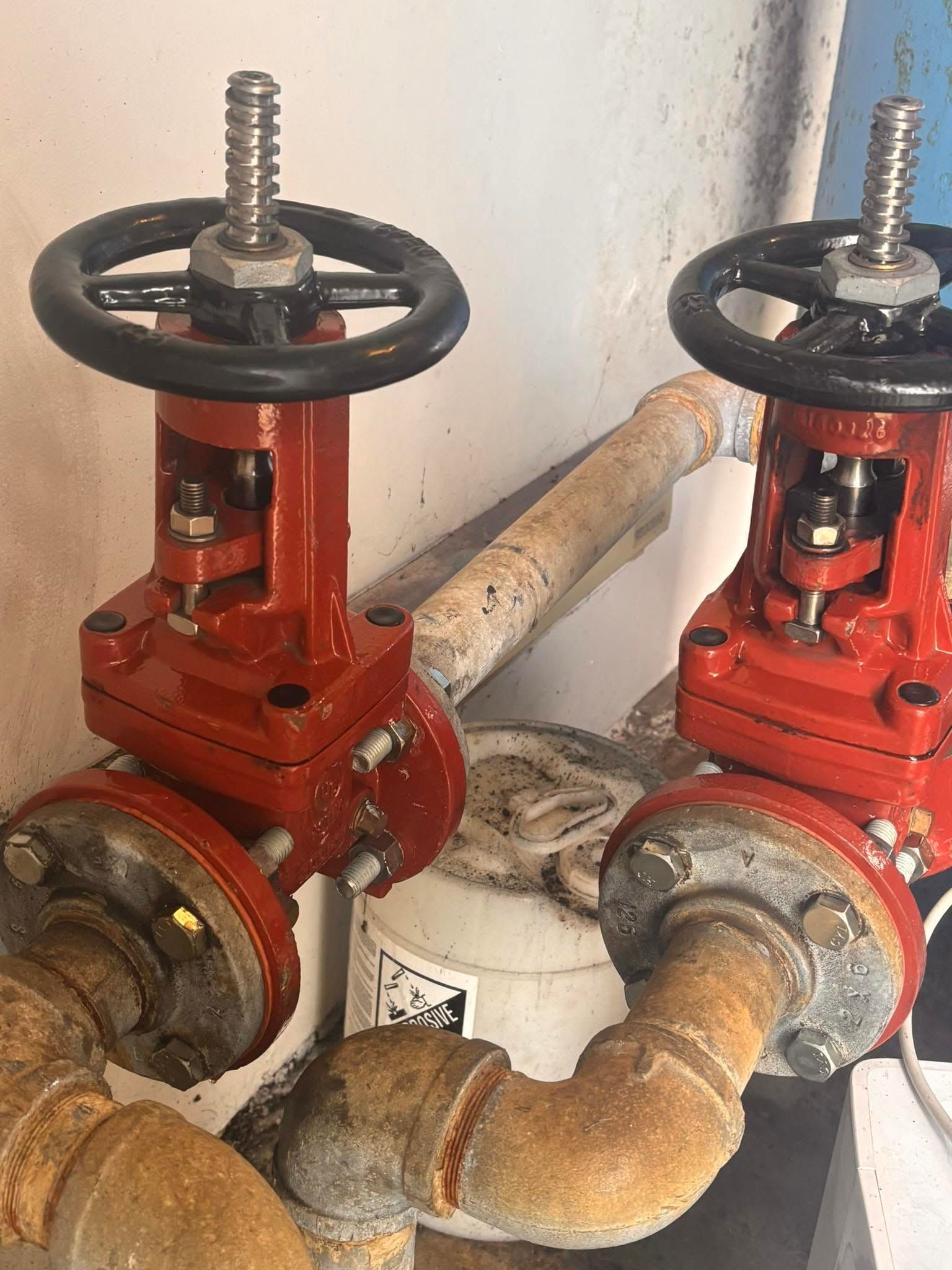 Two red industrial valves with black handwheels on rusted pipes against a white wall.