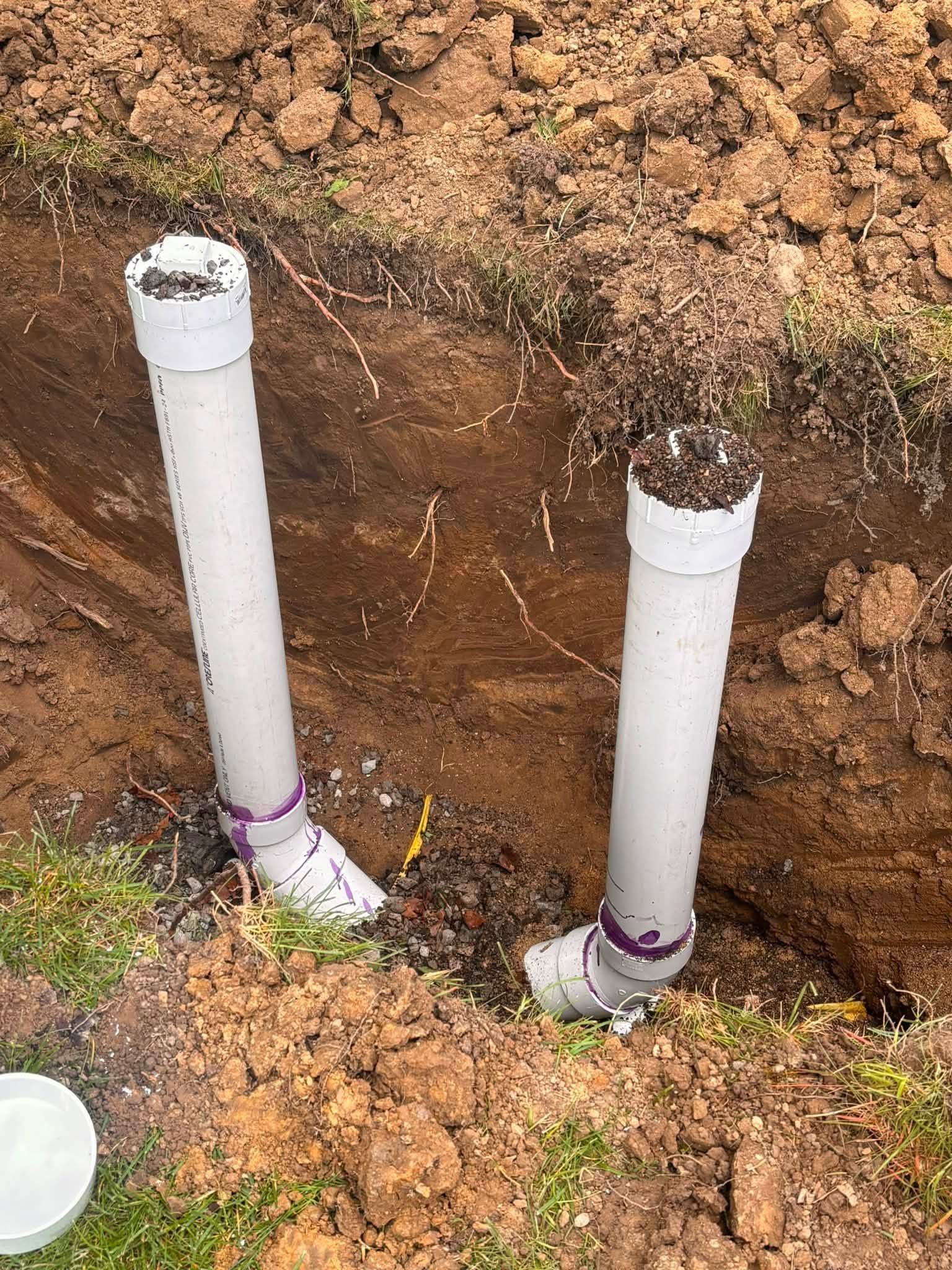 Two white PVC pipes standing in an excavated dirt trench, one capped and one open.
