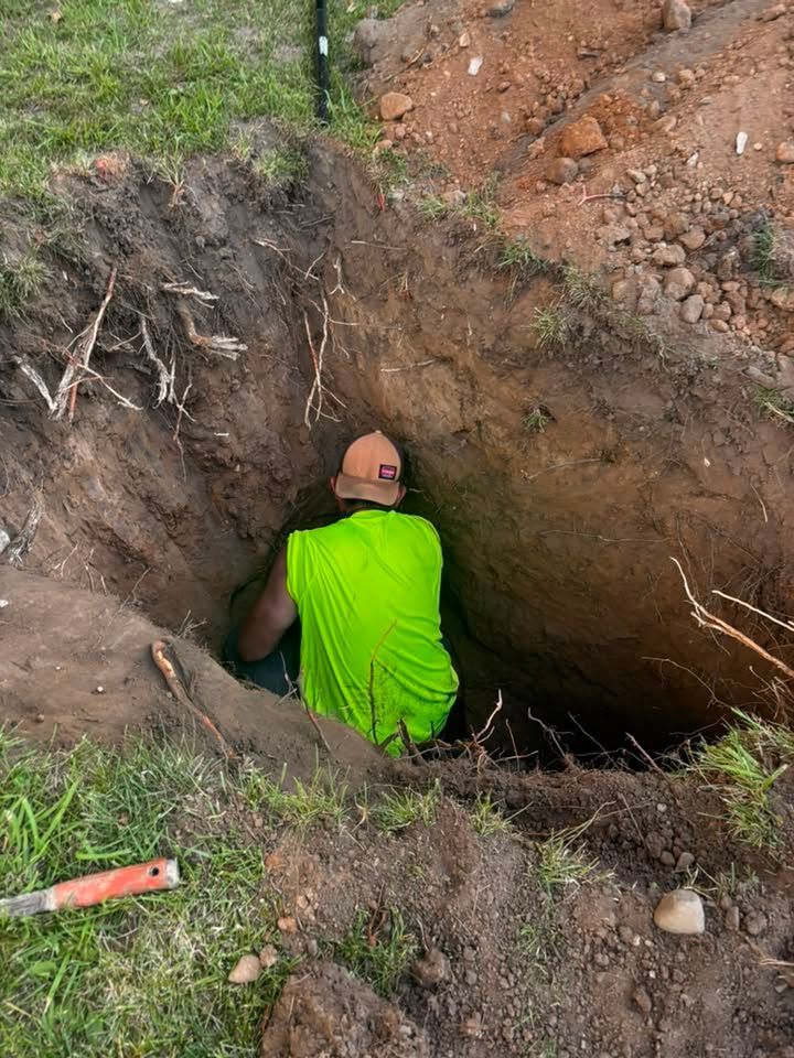 Worker in a neon green shirt and hard hat crouches in a deep dirt trench beside an excavation site.