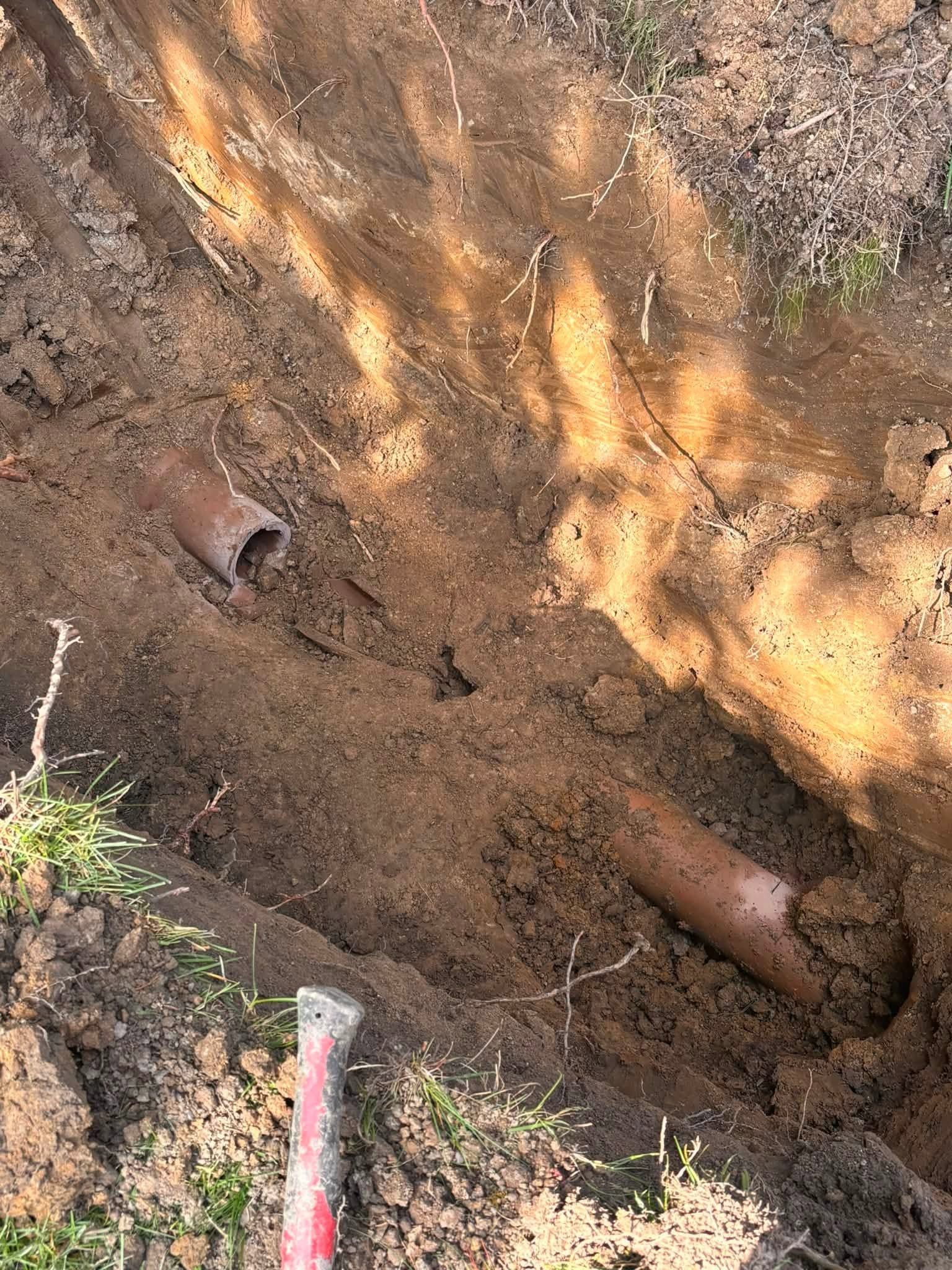 Rusty metal pipes exposed in a dirt trench, with soil and plant roots around them.